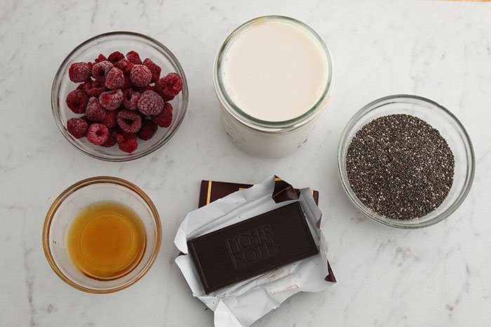 Raspberry Chocolate Chia Pudding raspberries, milk chia seeds, honey in bowls chocoalate in wrapper birds eye view on white marble