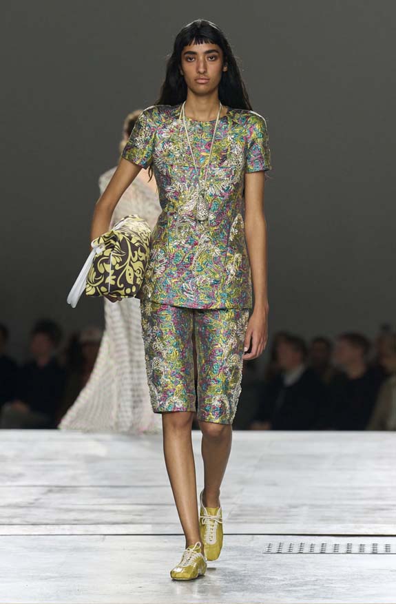 Dries Van Noten Spring 2026 runway showing gold metallic soft flat sneakers styled with a brocade walking shorts set