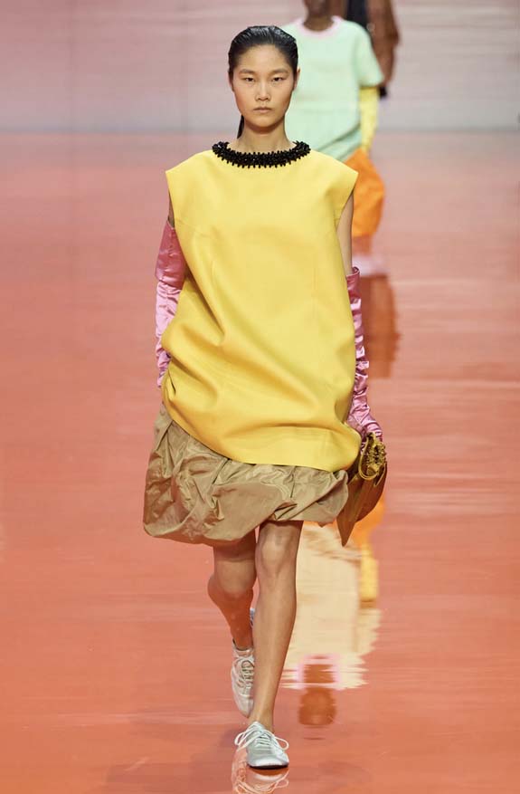 Prada Spring 2026 runway showing satin soft flat sneakers styled with a tunic and asymmetrical balloon midi skirt